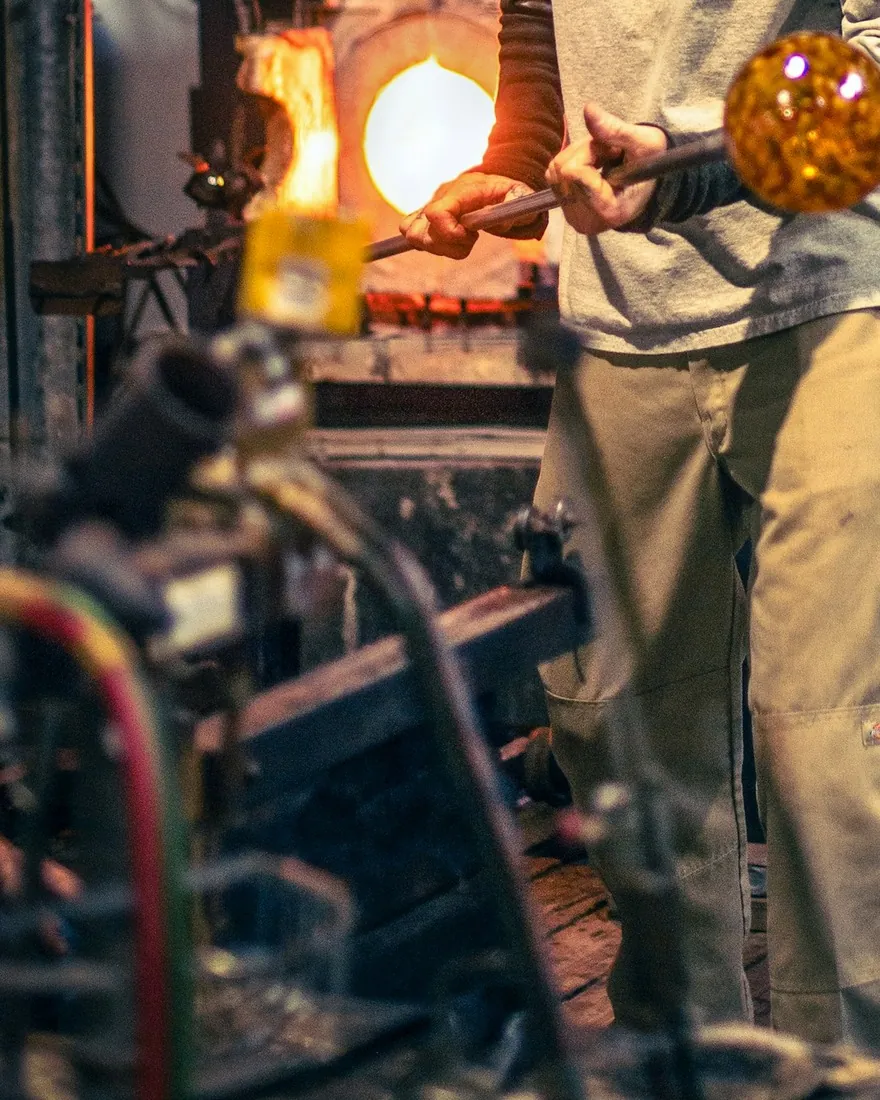 Manos sosteniendo la caña con una masa de vidrio incandescente ante la boca abierta del horno de recalentar